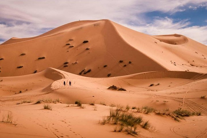 Two people walking below a high orange sand dune in Erg Chebbi
