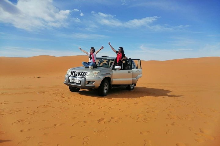 Two travelers posing with a stationary four-wheel-drive vehicle among orange dunes